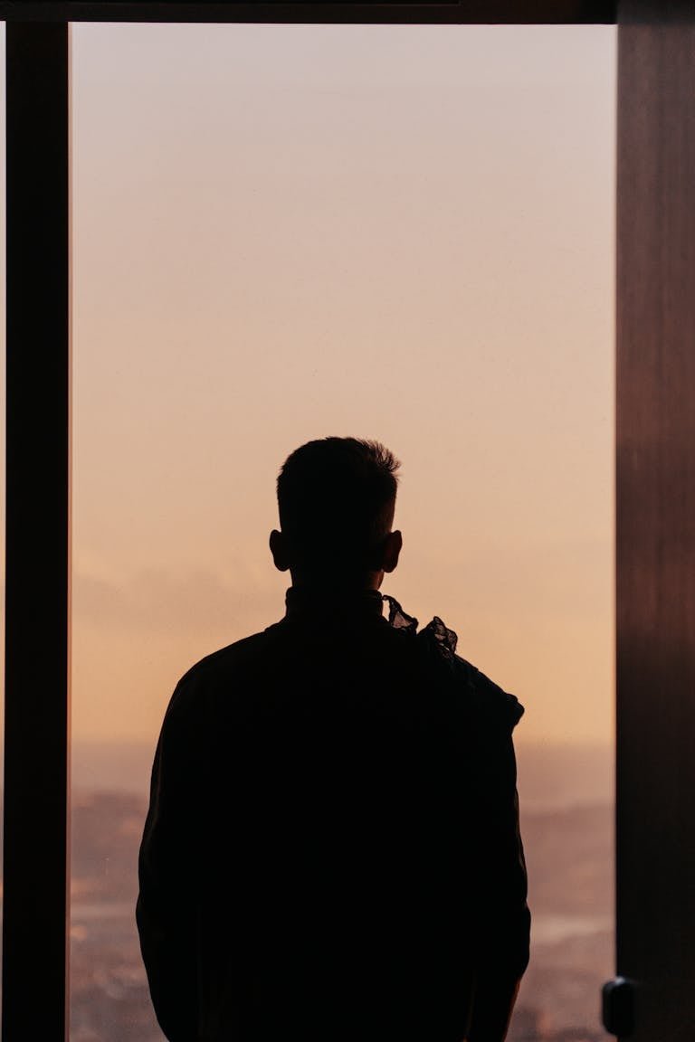 Silhouette of a man gazing out a window, capturing the serene sunset sky.