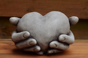 A stone sculpture of hands gently holding a heart against a wooden background, symbolizing protection and love.