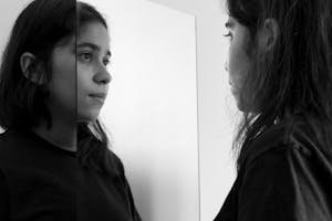 Black and white photo of a young woman reflecting on herself in a mirror indoors.